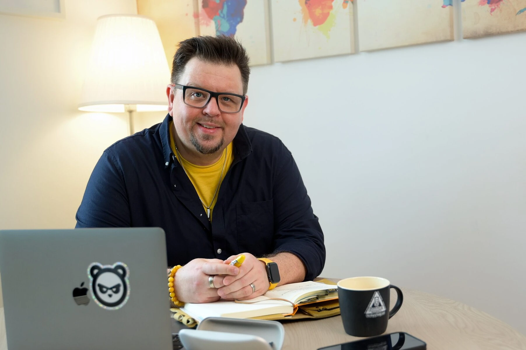 Kommunikationsprofi bei TheAngryTeddy Mann sitzt an Schreibtisch mit Laptop, Notizbuch und Tasse, arbeitet im Büro.